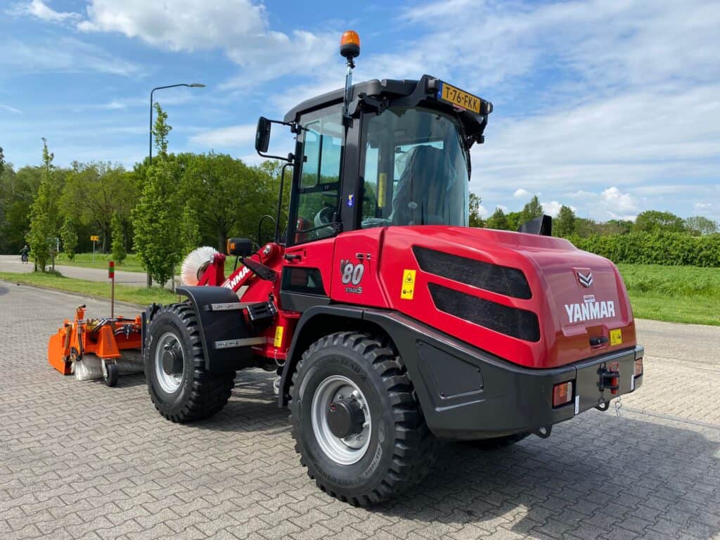 Stille landbouwwerktuig van Yanmar met achterborstel op bestrating met groene bomen en blauwe hemel in de achtergrond.