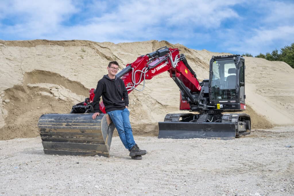 Mannelijke werknemer naast compacte graafmachine bij zandheuvels op bouwplaats.