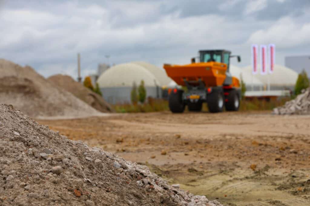Korte omschrijving van de bouwplaats met bulldozer en bouwmaterialen in de achtergrond.