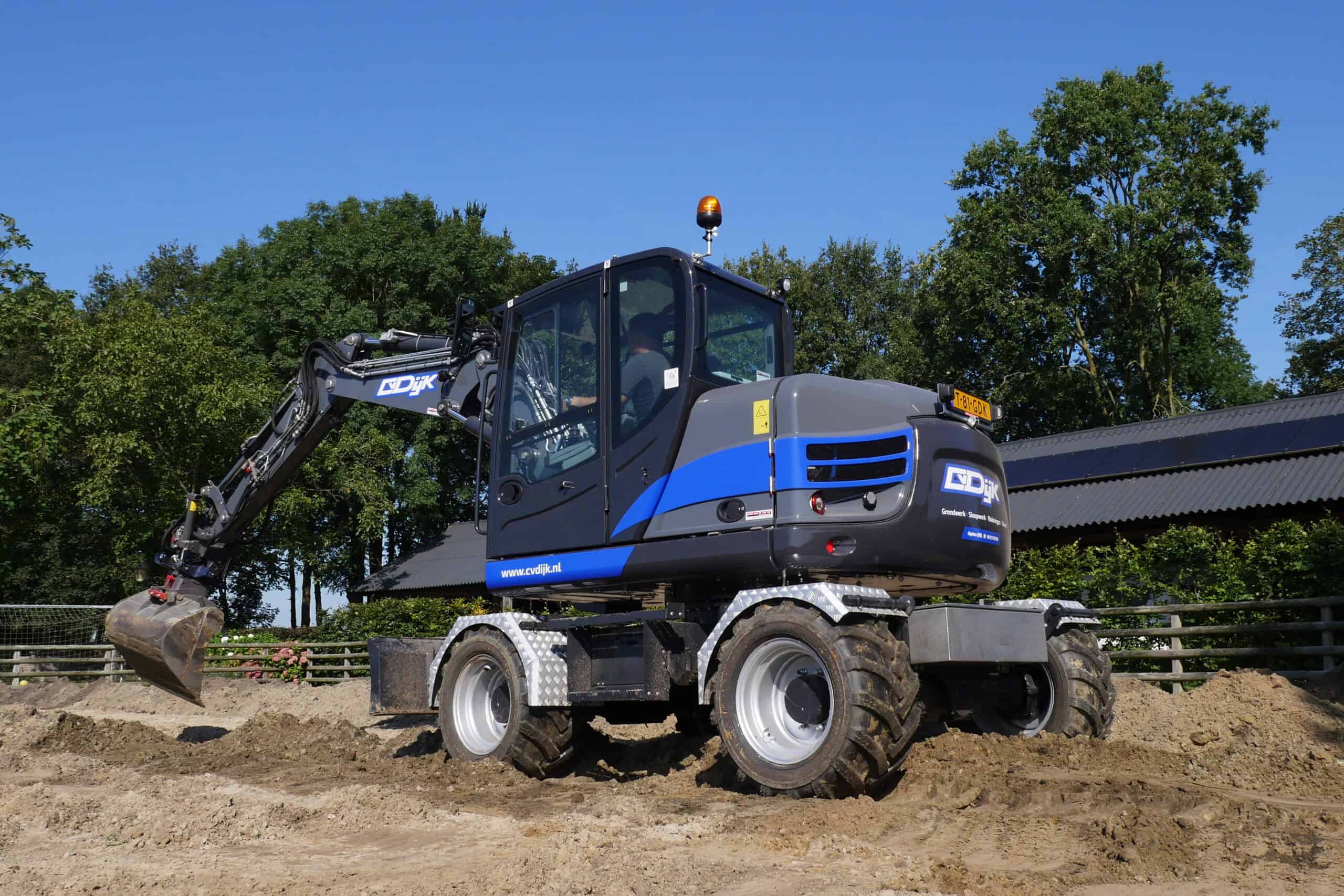Kleine robot graafmachine op bouwplaats met blauwe lucht en groene bomen.
